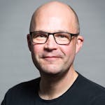 Portrait of a smiling bald man wearing glasses and a black t-shirt against a neutral background.