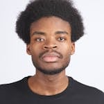 Professional studio portrait of a man with afro hair wearing a black t-shirt against a white backdrop.